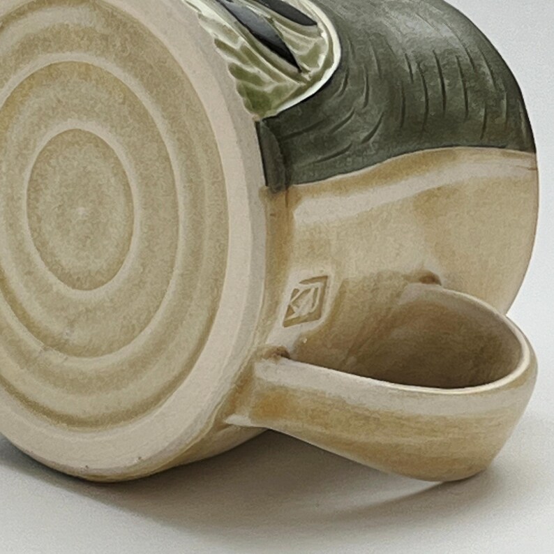 K&ouml;nnte beinhalten: Eine Keramik-Tasse mit beigefarbenem &Auml;u&szlig;eren und einem dunkelgr&uuml;nen und schwarzen Design. Die Tasse hat einen abgerundeten Henkel und einen kreisf&ouml;rmigen Boden mit konzentrischen Ringen. Ein kleines quadratisches Emblem ist in der N&auml;he des Henkels zu sehen.