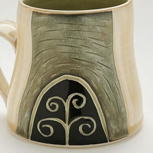 K&ouml;nnte beinhalten: Eine handgefertigte Keramiktasse mit cremefarbenem &Auml;u&szlig;eren und gr&uuml;nem Inneren. Die Tasse hat eine strukturierte, olivgr&uuml;ne Fl&auml;che mit einem schwarzen, bogenf&ouml;rmigen Design und einem wei&szlig;en Wirbelmuster. Der Griff ist cremefarben.
