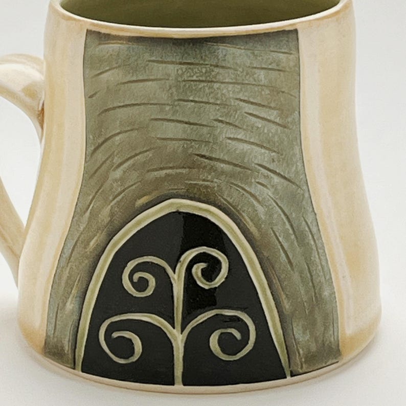 K&ouml;nnte beinhalten: Eine handgefertigte Keramiktasse mit cremefarbenem &Auml;u&szlig;eren und gr&uuml;nem Inneren. Die Tasse hat eine strukturierte, olivgr&uuml;ne Fl&auml;che mit einem schwarzen, bogenf&ouml;rmigen Design und einem wei&szlig;en Wirbelmuster. Der Griff ist cremefarben.
