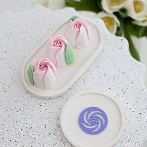 Puede incluir: Tres pasteles en forma de flor rosa y blanca en un plato ovalado blanco. Un plato redondo blanco con un diseño de espiral morado está sobre una superficie blanca moteada.