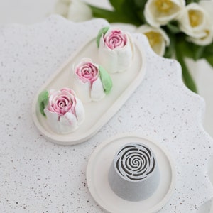 May include: Three white cake pops decorated with pink and white frosting roses on a white oval plate. A gray cake pop with a swirl design is on a white round plate.