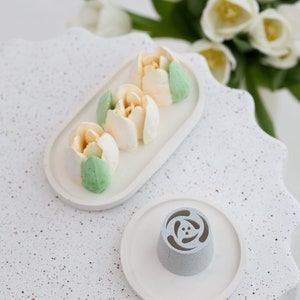 May include: A set of four white and green frosting tulips on a white oval plate. A white plate with a gray flower-shaped piping tip sits on a white speckled surface.