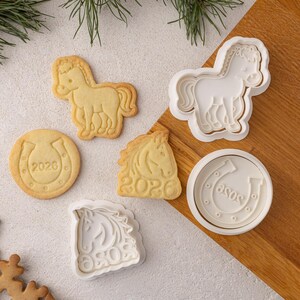 Puede incluir: Un conjunto de galletas con forma de caballo y cortadores de galletas. Las galletas son de color marrón claro y presentan un caballo, una herradura con el año 2026 y una cabeza de caballo. Los cortadores de galletas blancos tienen forma de caballo y herradura.