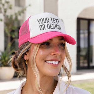 Puede incluir: Una gorra tipo camionero de color rosa brillante y blanco con un panel trasero de malla y un panel frontal rígido que presenta el texto YOUR TEXT OR DESIGN impreso en una fuente gris oscuro sin remates en la parte frontal.