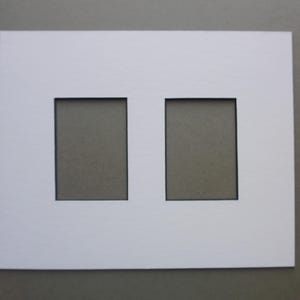 May include: A white picture mat with two rectangular openings. The mat is made of thick, bright white paper and is set against a neutral gray background. The openings are evenly spaced and have a black border.