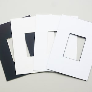 May include: Four different colored mat boards with a rectangular opening in the center. The mat boards are black, white, white, and white. The mat boards are stacked on top of each other.