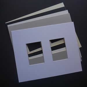 May include: A stack of various colored mat boards, including white, gray, and black. The top mat board is white with two rectangular openings. The mats are arranged on a dark surface, showcasing the different shades.