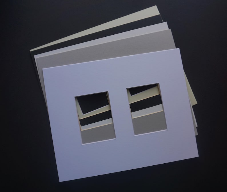 May include: A stack of various colored mat boards, including white, gray, and black. The top mat board is white with two rectangular openings. The mats are arranged on a dark surface, showcasing the different shades.