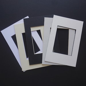 May include: A collection of rectangular picture mats in various colors, including white, cream, black, and gray. Each mat has a central rectangular opening. The mats are arranged on a dark surface, creating a contrast.