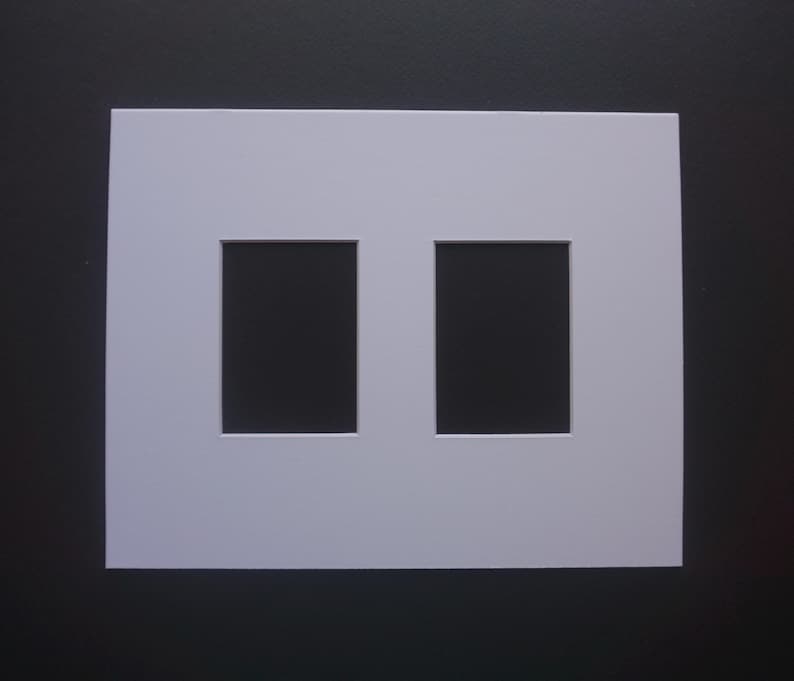 May include: A white picture mat with two rectangular openings. The mat is square and has a clean, minimalist design. The background is a dark gray color, providing contrast to the white mat.