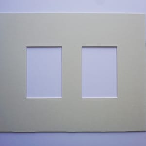 May include: A light green double picture mat with two rectangular openings. The mat is made of thick paper and has a clean, simple design. The openings are white, ready for photos or artwork.