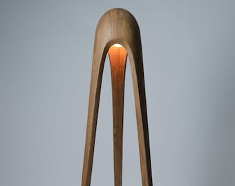 May include: A wooden floor lamp with a unique arched design. The lamp has three legs and a warm light emanating from the top. The wood has a natural finish, and the lamp is set against a neutral background.