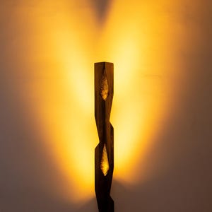May include: A dark brown wooden floor lamp with two illuminated, leaf-shaped cutouts. The lamp has a geometric design and stands on a square base. The light casts a warm glow on the wall behind it.