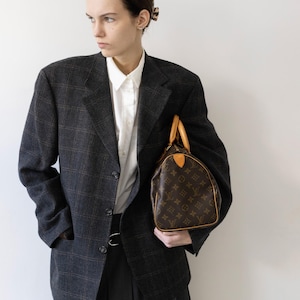 May include: A dark charcoal grey blazer with a subtle plaid pattern, worn over a crisp white collared shirt and a dark grey pleated skirt. A brown leather handbag with a repeating monogram print and tan leather handles is held at the side.