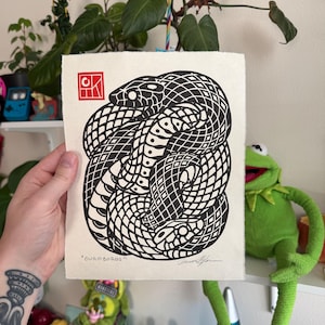 Può includere: Stampa in bianco e nero di un serpente avvolto a cerchio, con il testo "ouroboros" sotto. La stampa è su sfondo bianco e ha un timbro rosso nell'angolo in alto a sinistra.