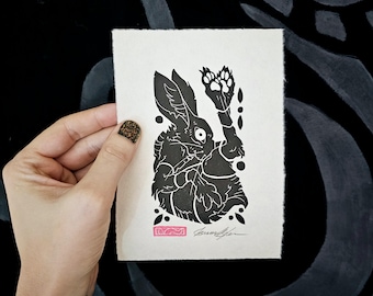 May include: Black and white print of a rabbit with one paw raised, on a white background with a pink signature.