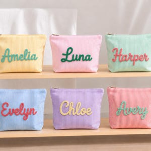 Puede incluir: Seis neceseres personalizados en colores pastel, cada uno con un nombre diferente bordado en un color contrastante. Las bolsas son a rayas y tienen cierre de cremallera. Los nombres son Amelia, Luna, Harper, Evelyn, Chloe y Avery.