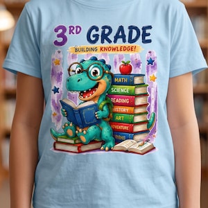 May include: A light blue t-shirt featuring a graphic of a green dinosaur wearing glasses and reading a book. The text reads 3RD GRADE BUILDING KNOWLEDGE. A stack of colorful books labeled Math, Science, Reading, History, Art, Adventure, and Rang is shown.