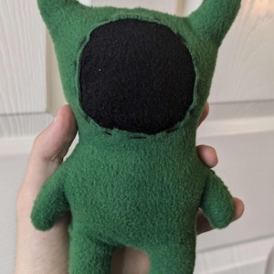 Puede incluir: Muñeco de peluche verde hecho a mano con una cara negra y dos cuernos. El muñeco tiene un diseño sencillo con detalles cosidos y brazos y piernas pequeños. El muñeco se sostiene en la mano de una persona.