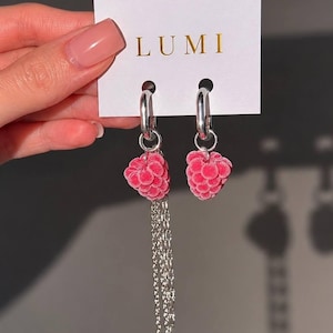 May include: A pair of silver hoop earrings featuring pink raspberry-shaped charms. One earring includes a long silver chain with a small white pearl and a white flower charm. The earrings are attached to a white display card with the text LUMI.