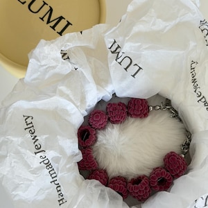 May include: A necklace featuring dark pink, textured, berry-shaped beads arranged in a circular pattern on a white faux fur base. The necklace is nestled inside white tissue paper printed with the text LUMI and Handmade Jewelry.