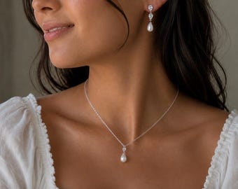 May include: A silver-toned necklace and matching drop earrings set. The necklace features a delicate chain with a single teardrop-shaped white pearl pendant accented by a small clear stone. The earrings mirror this design with a clear stone stud.