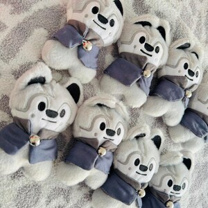 May include: A group of plush wolf toys. Each toy is white with grey details, black ears, and a smiling face. They are dressed in a blue and grey outfit with a small bell. The toys are displayed on a textured surface.