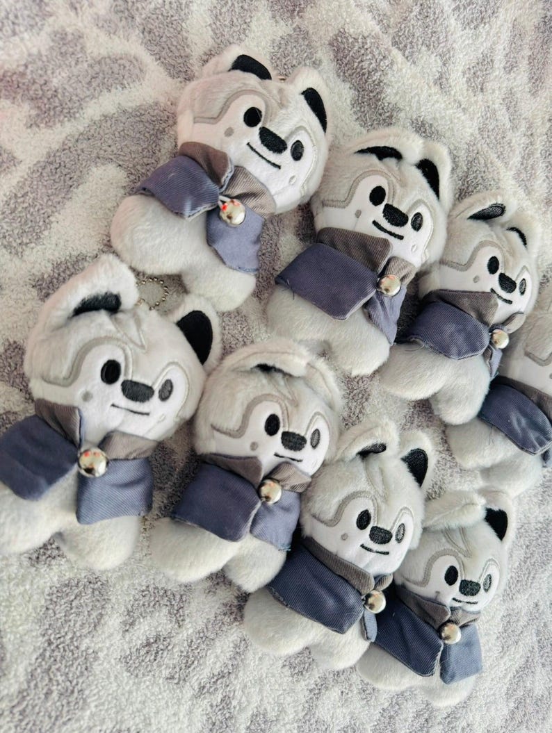 May include: A group of plush wolf toys. Each toy is white with grey details, black ears, and a smiling face. They are dressed in a blue and grey outfit with a small bell. The toys are displayed on a textured surface.