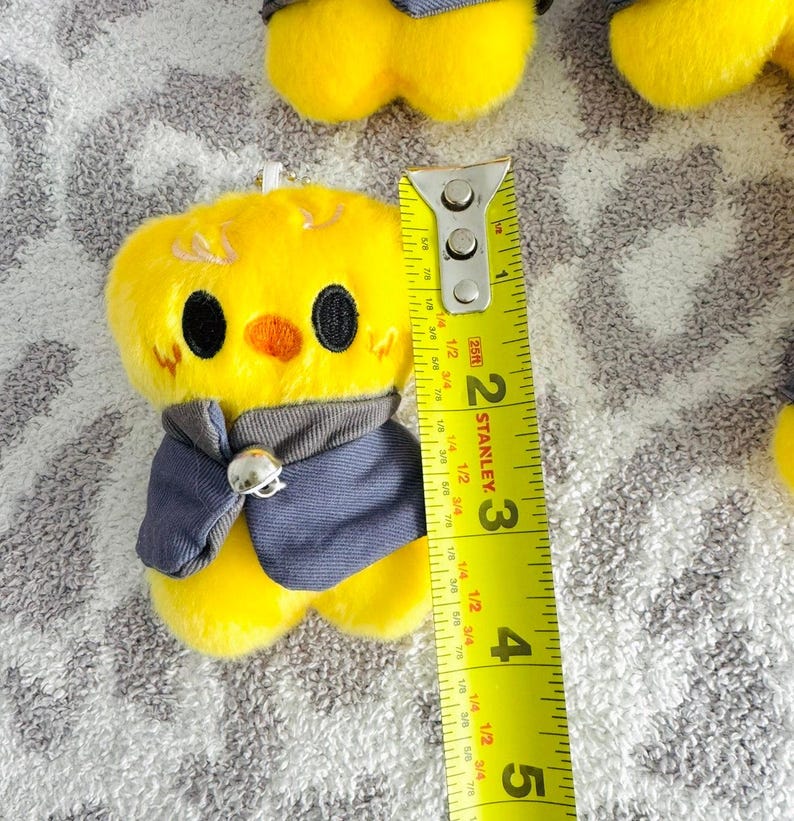 May include: A plush yellow chick with black eyes, an orange beak, and a grey vest. The chick is approximately 5 cm tall, as measured by a yellow and silver Stanley tape measure.