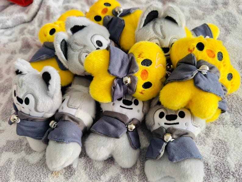 May include: A pile of plush toys in yellow and grey. The yellow toys have black accents, while the grey toys have black and white accents. Each toy is wearing a blue cape and a silver bell.