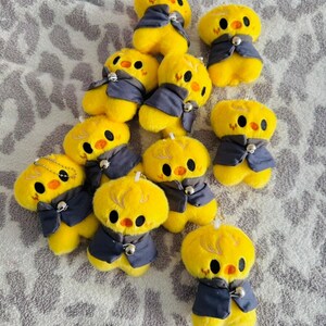 May include: A group of yellow chick plush toys, each dressed in a dark blue waistcoat with a silver bell. The chicks have black eyes and orange beaks. The toys are displayed on a patterned grey surface.