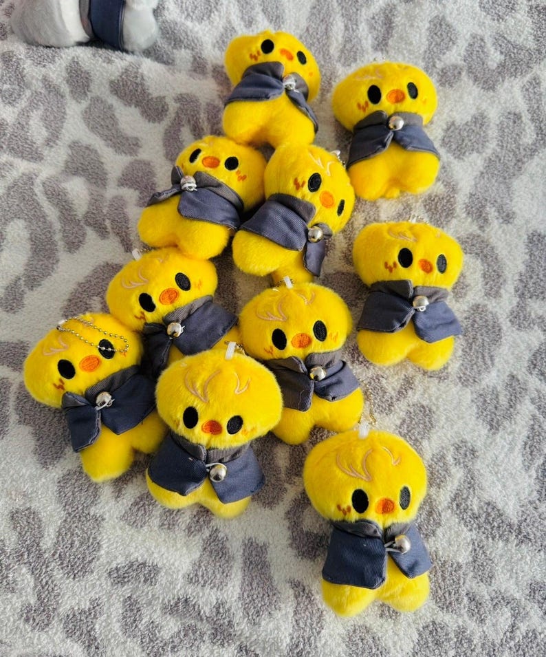May include: A group of yellow chick plush toys, each dressed in a dark blue waistcoat with a silver bell. The chicks have black eyes and orange beaks. The toys are displayed on a patterned grey surface.
