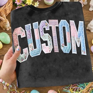 May include: Black sweatshirt with the word "CUSTOM" appliqued across the front in a patchwork design. The letters are made of various pastel-colored fabrics, including floral and gingham patterns. The sweatshirt is surrounded by Easter decorations.