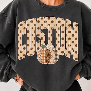 Pode incluir: Uma camisola de gola redonda cinzento-carvão com a palavra CUSTOM em letras grandes bege com bolinhas. Abaixo do texto, há um gráfico de uma abóbora com segmentos que exibem vários padrões, incluindo xadrez, estampa de leopardo e flores.