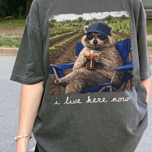 May include: Dark grey t-shirt featuring a raccoon wearing sunglasses and a cap, relaxing in a blue chair with a drink. The graphic includes a field scene and the text "i live here now".