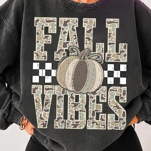 May include: A charcoal grey crewneck sweatshirt featuring a large graphic print with the text FALL VIBES in a camouflage pattern. The design includes a central pumpkin illustration and a checkered border element integrated into the lettering.