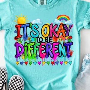 Puede incluir: Una camiseta azul claro con el texto "It's Okay to Be Different" en letras coloridas estilo grafiti. La camiseta presenta un sol, un arcoíris, nubes, flores, piezas de rompecabezas y huellas de manos. También se ven un par de zapatos sin cordones a cuadros blancos y negros y unos vaqueros.
