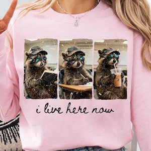 Op de afbeelding: Roze sweatshirt met een afbeelding van drie wasberen met hoeden en zonnebrillen in een klaslokaal. De wasberen worden afgebeeld terwijl ze lezen, schrijven en koffie drinken. De tekst "i live here now" staat eronder.