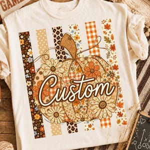 Peut inclure: Un t-shirt couleur crème arborant un grand motif central représentant une citrouille décorée de divers motifs floraux, léopard et à carreaux. Le mot Custom est écrit en lettres cursives marron sur la citrouille, devant des rayures verticales.