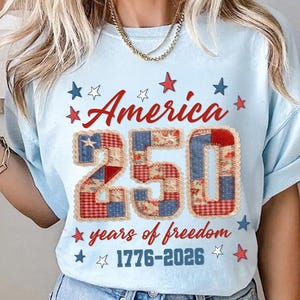 May include: Light blue t-shirt with the text "America 250 years of freedom 1776-2026". The number 250 is designed with a patchwork pattern in red, white, and blue. Red and blue stars surround the text.
