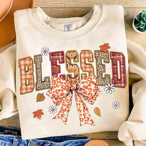 May include: A cream-colored sweatshirt with the word "Blessed" in orange, red, and brown plaid, leopard print, and floral patterns. A large orange and brown bow with leopard print accents is centered below the word.