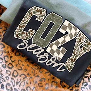 May include: A folded black sweatshirt with the words "COZY Season" in a distressed, layered design. The letters feature camouflage and checkered patterns. Additional folded shirts in shades of green and blue are visible in the background.
