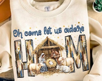 May include: A beige sweatshirt with a Christmas nativity scene design. The design features a brown wooden stable with a thatched roof, a manger with a baby Jesus, Mary, Joseph, and two sheep. The text "Oh come let us adore HIM" is written in blue and white letters above the scene.