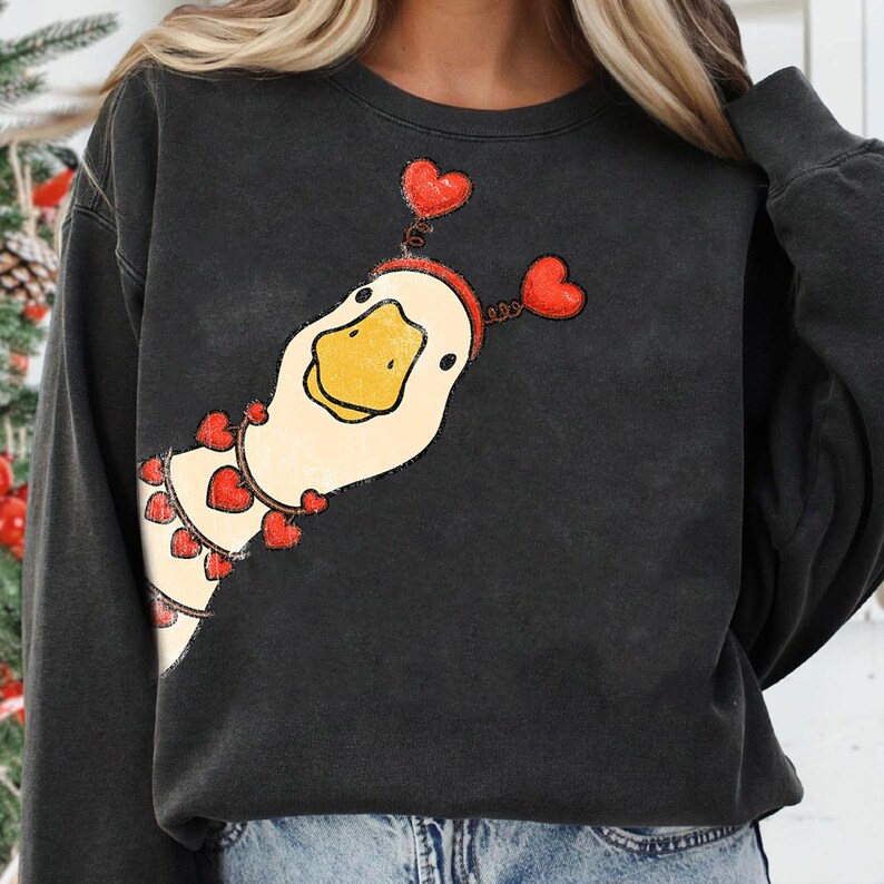 May include: Dark gray sweatshirt featuring a cartoon goose with a yellow beak and a red heart-shaped headband. The goose is also wearing a necklace of red hearts. The sweatshirt is a casual style.