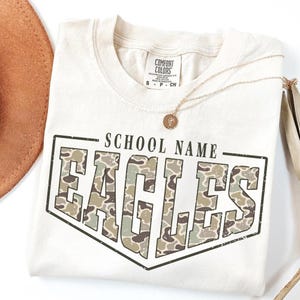 May include: An off-white short-sleeve t-shirt featuring the text SCHOOL NAME above the word EAGLES in large block letters. The letters are filled with a green, brown, and tan camouflage pattern, all enclosed within a dark outline border.
