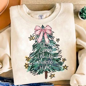 May include: A cream-colored sweatshirt with a watercolor Christmas tree design. The tree has a pink bow and the words "Oh Come Let Us Adore Him" written on it.