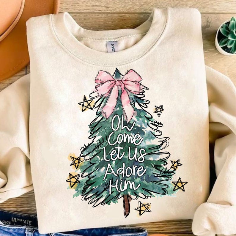 May include: A cream-colored sweatshirt with a watercolor Christmas tree design. The tree has a pink bow and the words "Oh Come Let Us Adore Him" written on it.
