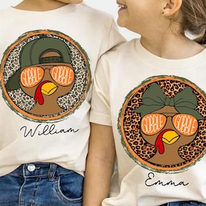 May include: Two off-white t-shirts with turkey graphics. One shirt features a turkey wearing a green cap and orange sunglasses with "GOBBLE GOBBLE" on the lenses, and the name "William" below. The other shirt has a turkey wearing a green bow and orange sunglasses with "GOBBLE GOBBLE" on the lenses, and the name "Emma" below.