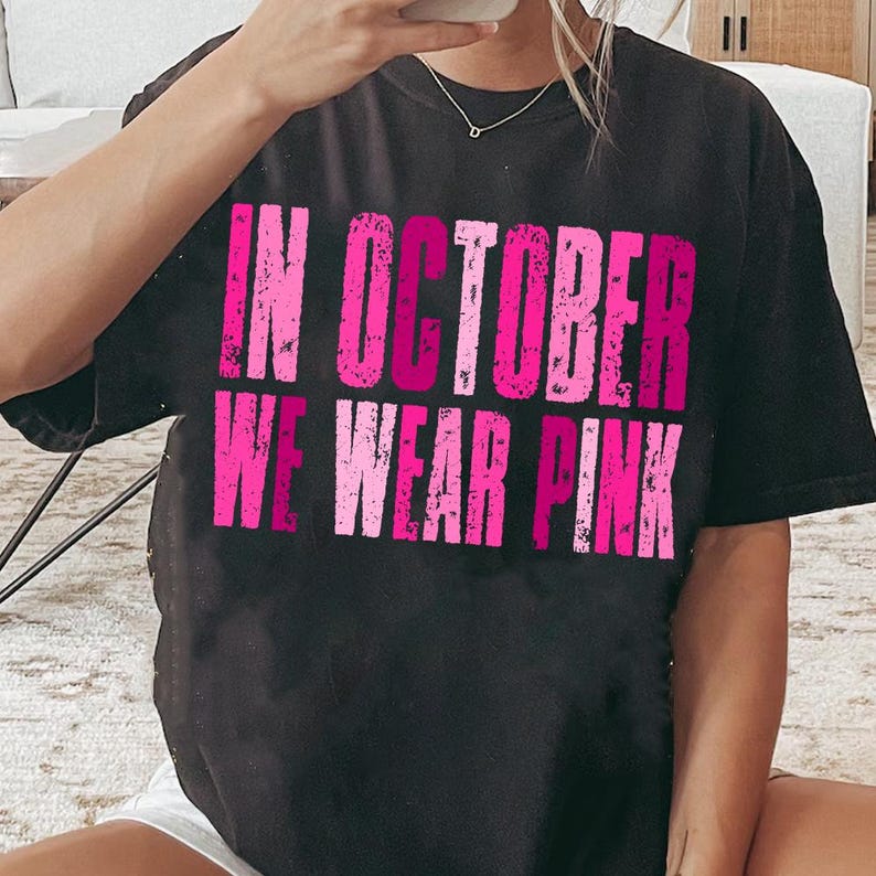 May include: Black t-shirt with the text "IN OCTOBER WE WEAR PINK" in distressed pink lettering. The shirt is a crew neck style and appears to be made of a soft material. The text is in a bold, sans-serif font.