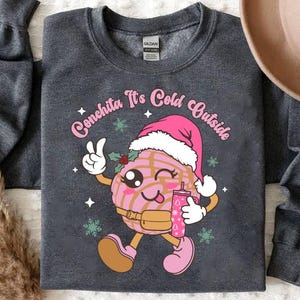 May include: A dark gray sweatshirt with a pink and brown cartoon concha wearing a Santa hat and holding a cup. The text "Conchita It's Cold Outside" is printed on the sweatshirt.